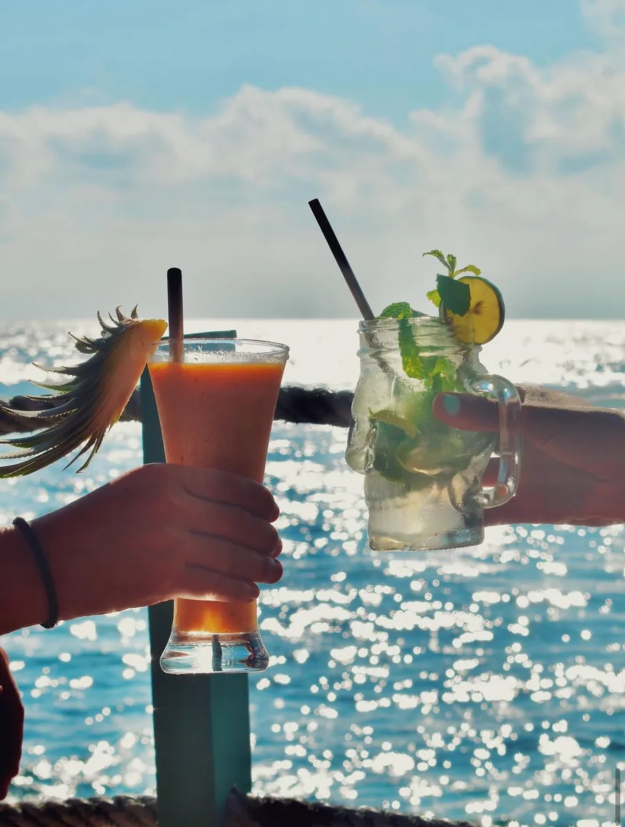 Two hands raising a mango juice and a mint drink served in a skull-shaped glass jar, against the sunlit sea.