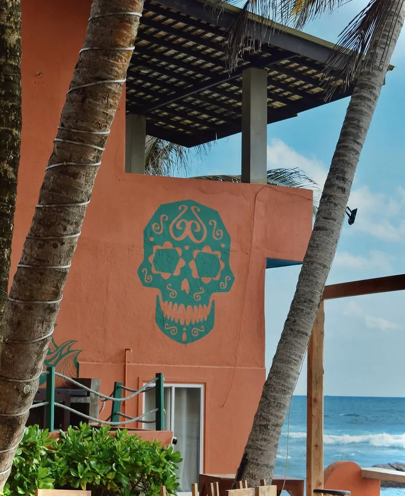 The ornamented skull painted in turquoise on the terracotta wall of the building, framed by two palm trunks, with the sea behind.