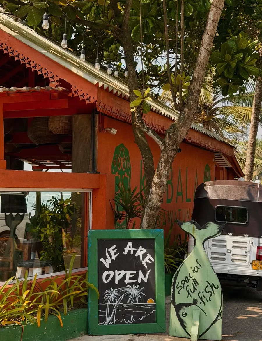 The entrance from the road: a chalkboard reading We Are Open, a fish-shaped sign, and a tuk-tuk parked by the terracotta wall.