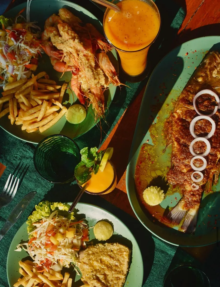 Overhead view of a full table: prawns with chips, a whole grilled fish topped with onion rings, salads and mango juices.
