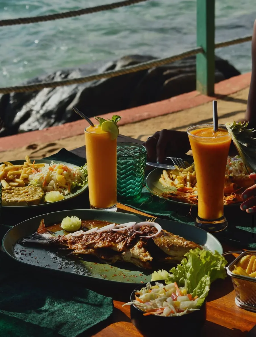 A grilled fish, prawns and two mango juices on a green cloth, with the rocks and the water a step away.