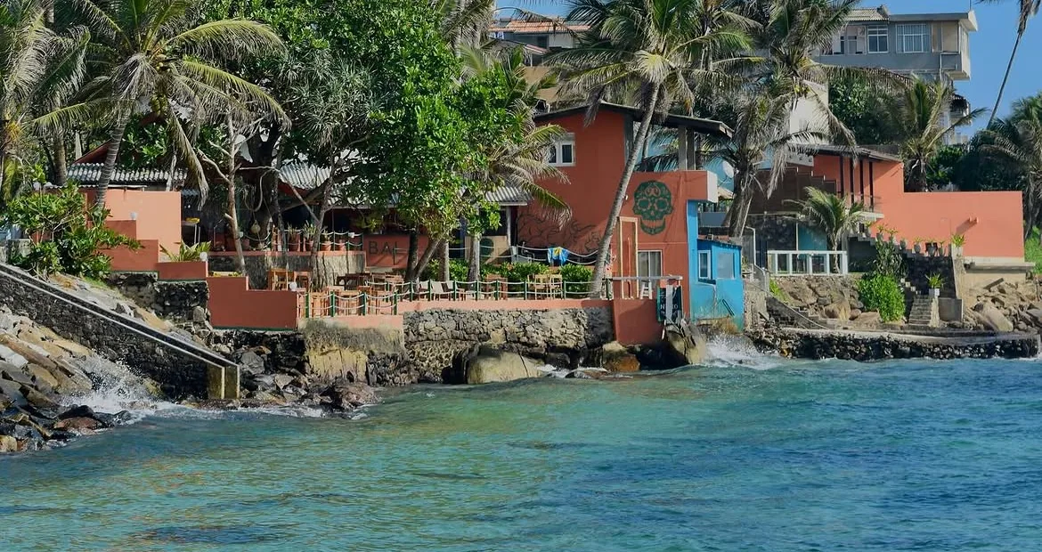 Kabala seen from the water: terracotta buildings and a sand terrace on the rocks, coconut palms above, turquoise sea in front.