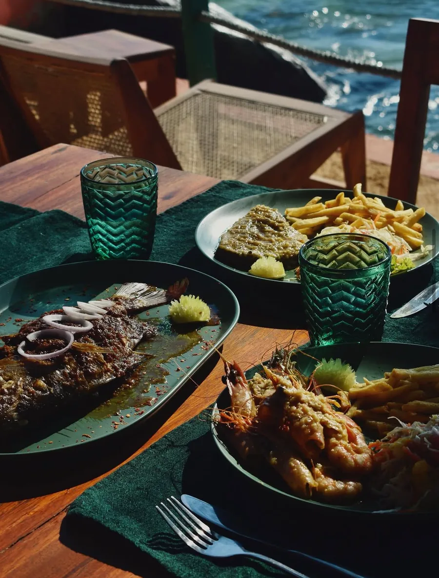 A whole grilled fish and a plate of prawns, both with chips and salad, on a table by the water.