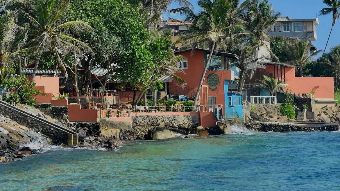 Kabala from the water: the sand terrace and the terracotta building with the skull on its wall, coconut palms above, turquoise sea in front.